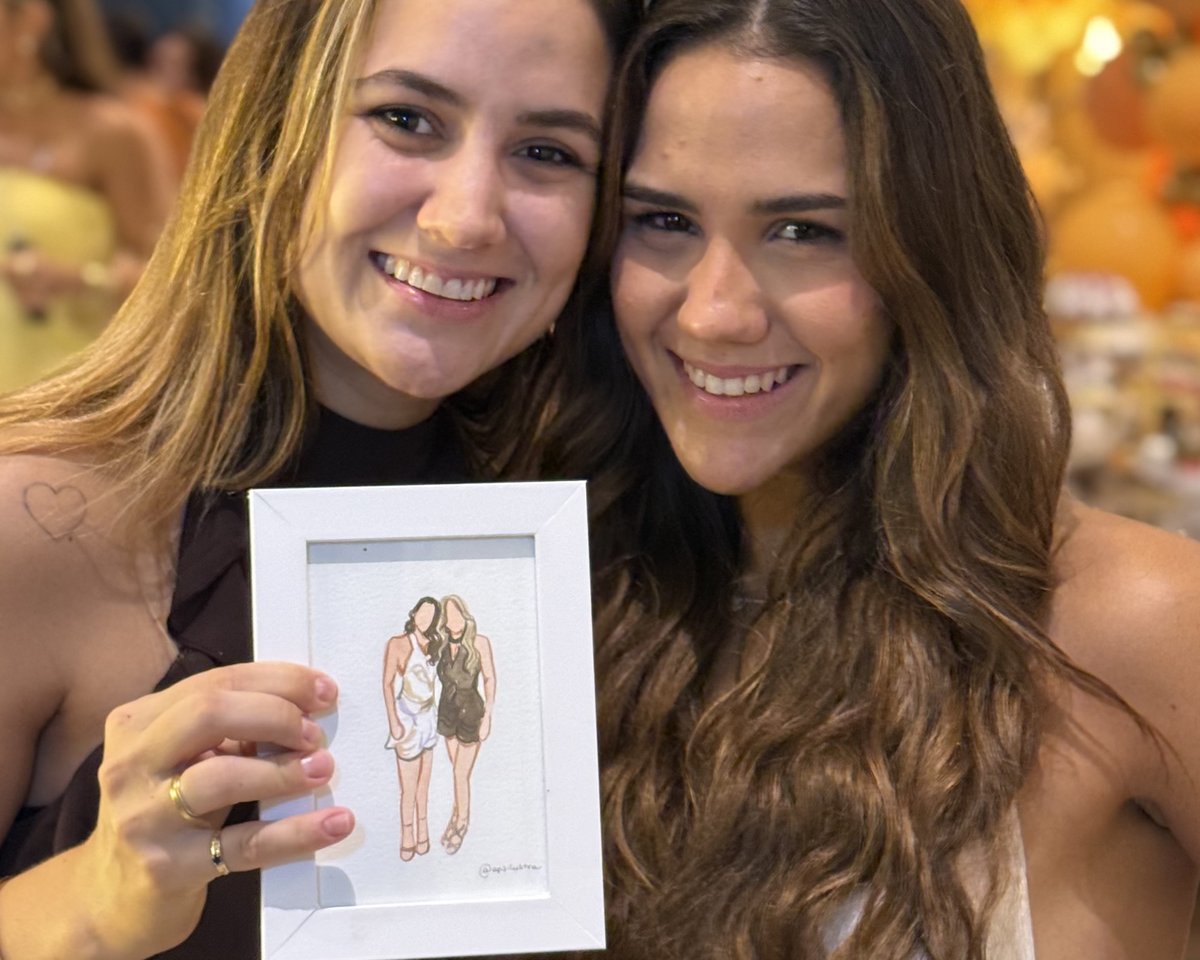 Duas convidadas sorrindo com o retrato delas em aquarela, na moldura