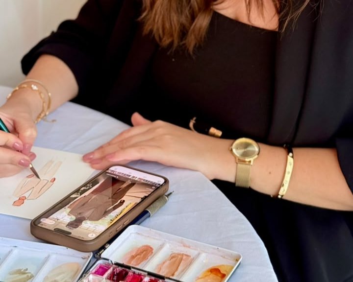 Mão da Ana pintando o esboço de um convidado, com o celular mostrando a foto de referência e a paleta de aquarela ao lado